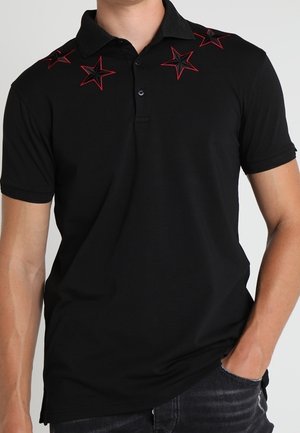 Sort polo t-shirt med korte ærmer og rød og sort stjernebroderi på skuldrene, båret af en person med den ene hånd i lommen.