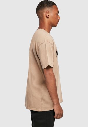 Homme portant un T-shirt beige surdimensionné et un pantalon noir, debout de profil sur un fond gris clair uni.