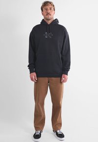 Sweat à capuche en coton noir avec poche frontale et motif graphique bleu, associé à un pantalon marron et des baskets noires et blanches.