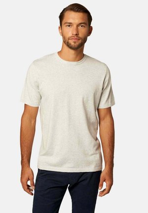 Mann mit kurzen braunen Haaren und Bart, der ein schlichtes hellgraues T-Shirt mit kurzen Ärmeln und dunkle Hosen trägt, vor weißem Hintergrund stehend.