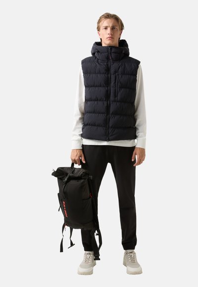 Námořnická pufovaná vesta s kapucí a zipem, v kombinaci s černými kalhotami. Model drží černý batoh s rolovacím vrchem a červenými akcenty.