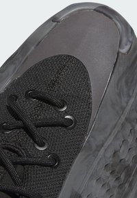 Fekete sportcipő, amelynek textúrázott hálós felsőrésze, fényes szürke díszítései és fekete fűzői vannak, amelyek kör alakú fűzőlyukakon futnak keresztül. A talprész gumi, amely jól látható.