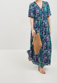 Sukienka maxi w kolorze zielonym z wzorem paisley, krótkie rękawy, głęboki dekolt w serek, taliowana i z lejącym materiałem. W skład zestawu wchodzi także torba pleciona oraz sandały na ramiączkach.