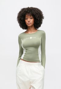 Γυναίκα με σγουρά μαλλιά που φοράει εφαρμοστή μακρυμάνικη μπλούζα σε ελιά πράσινο χρώμα με το κείμενο "Nike Shox" και λευκή φαρδιά παντελόνα, με τα χέρια στις τσέπες, ουδέτερο φόντο.