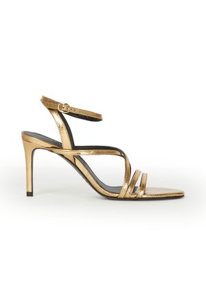Sandale à talons hauts métallisée dorée avec fine bride cheville, orteils découverts et design élégant à brides multiples sur fond blanc.