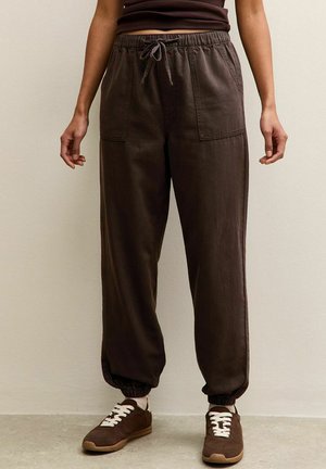 Persona che indossa pantaloni marrone scuro con coulisse e polsini elastici e sneakers marroni abbinate con lacci bianchi, in piedi contro un muro beige.