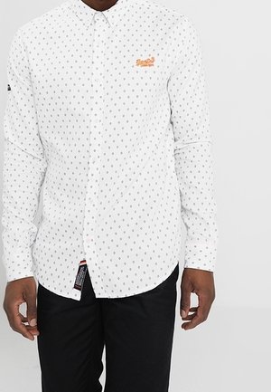 Man wearing white button-up shirt with small black diamond pattern and orange embroidered logo on chest, paired with black pants.