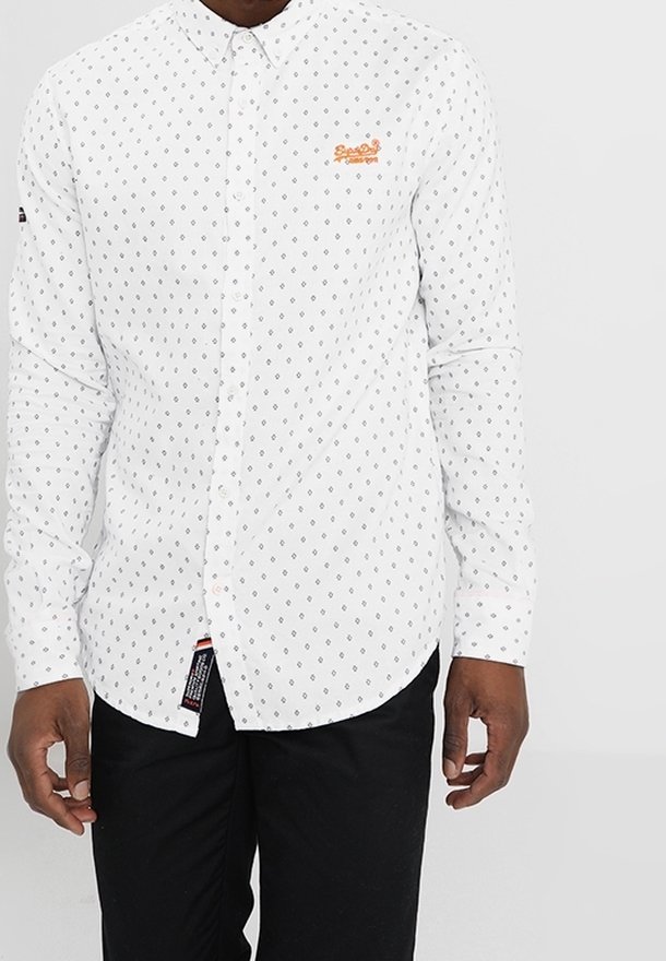 Homme portant une chemise blanche à boutons avec un petit motif de losanges noirs et un logo brodé orange sur la poitrine, associée à un pantalon noir.