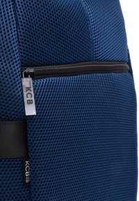 Mochila de malla azul con un bolsillo con cremallera y una correa negra. Etiqueta del logo de KCB visible. Superficie texturizada con un patrón de panal.