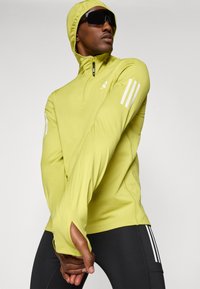 Gelbe zip-up Jacke mit langen Ärmeln und Kapuze, mit Daumenlöchern, weißen Streifenakzenten an den Ärmeln und einer glatten, dehnbaren Textur.