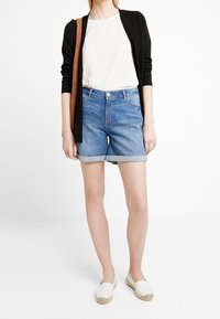 Pantalones cortos de mezclilla con lavado azul claro, dobladillos doblados, bolsillos frontales, combinados con una camiseta blanca y una chaqueta negra. Zapatos espadrilles blancos.