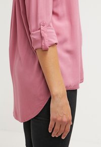 Chemise rose poudré à manches longues, texture soyeuse, poignets boutonnés, ourlet arrondi, associée à un pantalon noir. Accent sur les détails du tissu et de la coupe.