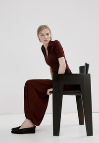 Woman in maroon ribbed top and loose pants sitting sideways on a black modern chair in a minimalist white room, looking forward.