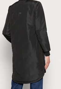 Personne portant une longue doudoune noire avec des poignets côtelés, un détail de fermeture éclair latérale, et un jean bleu, vue de dos.