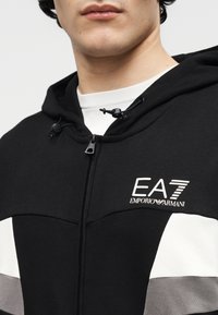 Muž v čiernej mikine na zips s kapucňou s bielymi a sivými pásmi a bielym logom EA7 Emporio Armani na hrudi.