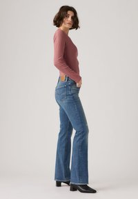 Γυναίκα με εφαρμοστό μαβί μακρυμάνικο τοπ, τζιν καμπάνα ψηλόμεσο σε μπλε χρώμα, μαύρα μπότες με τακούνι, στέκεται πλαγίως με το ένα χέρι στην τσέπη.