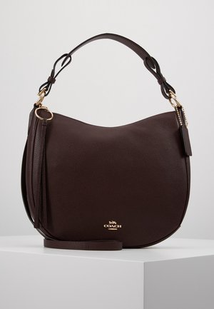 Sac à main Coach en cuir grainé marron foncé avec garnitures dorées, poignée supérieure unique, détail pompon et logo embossé sur le devant, exposé sur un piédestal blanc.