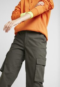 Henkilö, jolla on päällään oranssi huppari ja oliivinvihreät cargo-housut sivutaskuilla, rullaa yhtä hihaa ylös neonkeltaisen pitkähihaisen paidan yli.