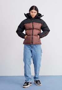 Chaqueta acolchada marrón y negra con capucha, con diseño acolchado y un emblema, combinada con jeans de mezclilla azul claros y zapatillas negras.