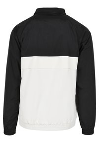 Giacca bianca e nera con colletto alto, polsini elasticizzati e una striscia bianca orizzontale sulla parte superiore della schiena. Realizzata in un materiale liscio e leggero.