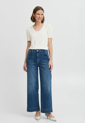 Top bianco a maniche corte con bordi texturizzati, abbinato a jeans in denim blu a vita alta e gamba larga, dotati di tasche a toppa frontali e orli arrotolati.