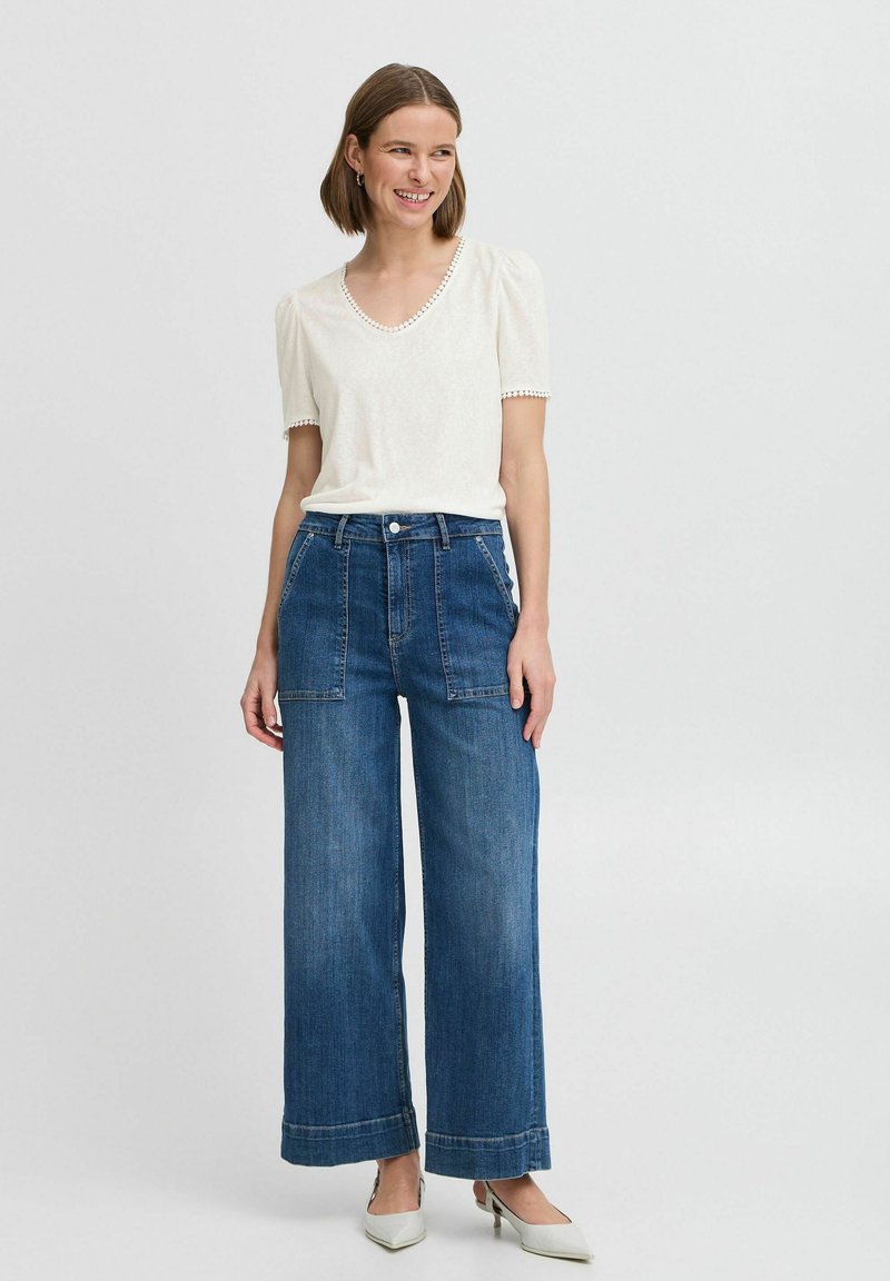 Haut blanc à manches courtes avec un bord texturé, associé à un jean en denim bleu à taille haute et à jambes larges, avec des poches plaquées à l'avant et des ourlets retroussés.