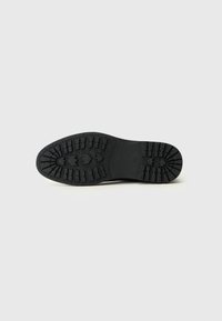 Semelle extérieure en caoutchouc noir avec un motif de semelle profond, présentant une surface texturée et des crêtes pour une meilleure adhérence. Logo circulaire situé près du talon.