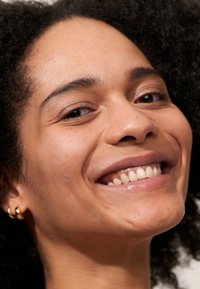Gros plan sur un visage souriant avec une peau marron moyen, des cheveux bouclés, des boucles d'oreilles en or, et des patchs circulaires clairs sur les joues.