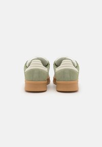 Zapatillas de ante verde con un panel de cuero crema, que presentan una suela de goma texturizada y un cuello acolchado. La vista trasera muestra detalles de costura simples.