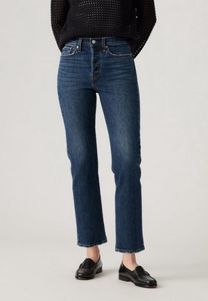 Personne portant un jean droit bleu, un pull en tricot noir et des mocassins en cuir noir, debout sur un fond uni.