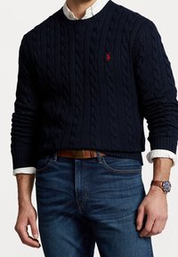 Marinblå kabelstickad tröja med rund hals och rött märke. Bärs med blå jeans och ett brunt läderbälte, visar en klocka på handleden.