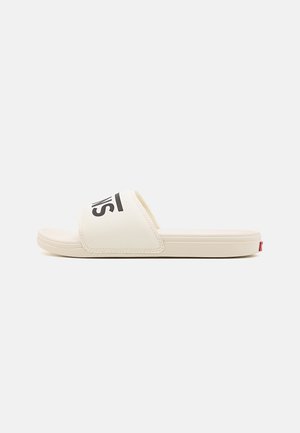Slide sandal in cream with a wide strap and black logo. Features a flat rubber sole and noticeable stitch detailing on the strap.