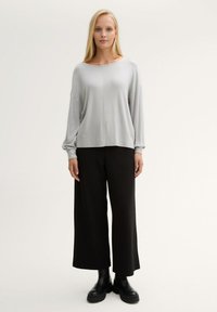 Hellgraues langärmliges Oberteil mit weitem Ausschnitt, kombiniert mit schwarzen, weiten Hosen. Das Modell trägt klobige schwarze Stiefeletten. Glatte, weiche Texturen.