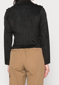 Veste noire en suédine synthétique avec un col structuré et des épaulettes ; design court. Portée avec un pantalon beige, mettant en valeur une texture lisse.