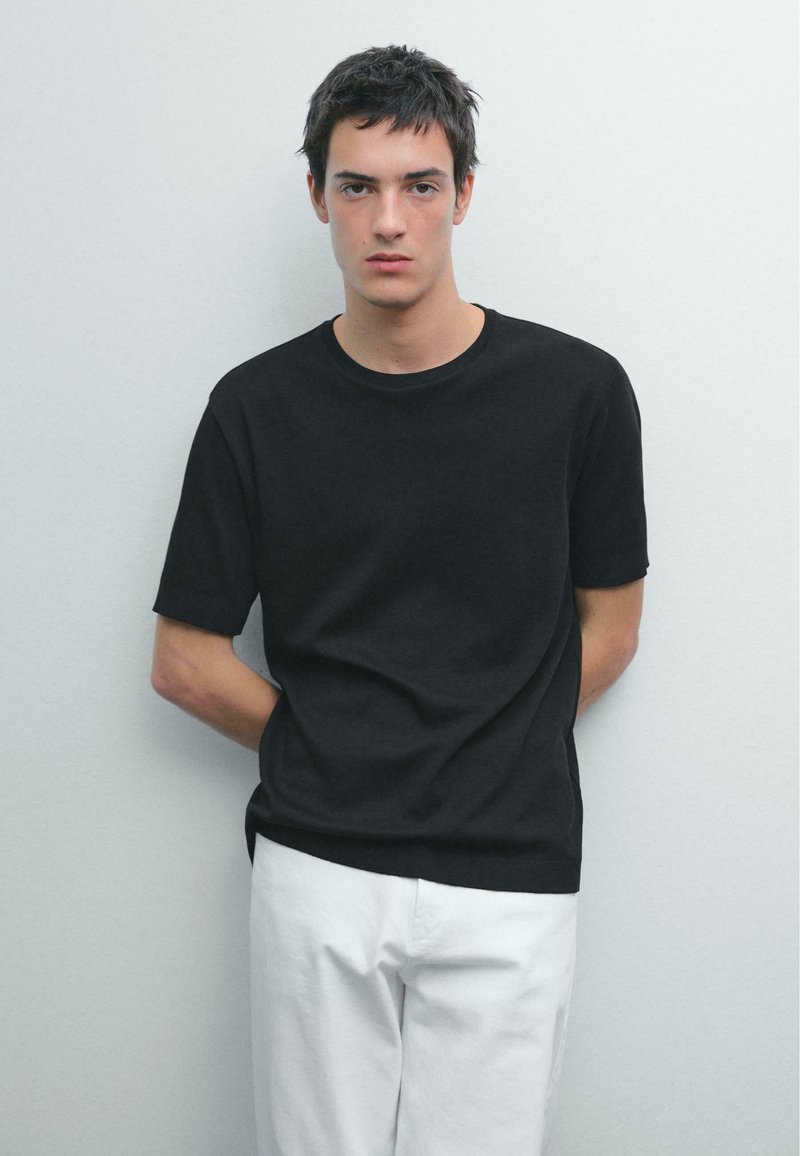 T-shirt nera a girocollo realizzata in tessuto morbido, maniche corte, con vestibilità comoda, abbinata a pantaloni bianchi. Design semplice e minimalista.