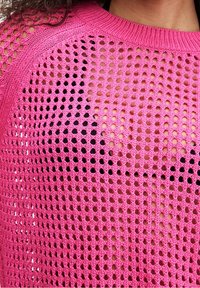 Różowy dzianinowy sweter z luźnym, otwartym splotem, okrągłym dekoltem oraz naprzemiennymi dużymi i małymi wzorami dziur.