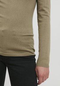 Suéter de manga larga verde olivo con un diseño ajustado, puños acanalados y textura suave, combinado con pantalones negros.