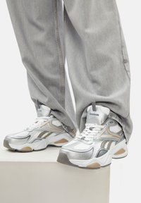 Persona ar brīviem gaiši pelēkiem džinsiem un baltiem Reebok sporta apaviem ar sudraba un bēša akcentiem, stāv uz baltas platformas.