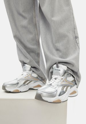 Osoba v voľných svetlosivých džínsoch a bielych teniskách Reebok so striebornými a béžovými akcentmi, stojaca na bielom podstavci.