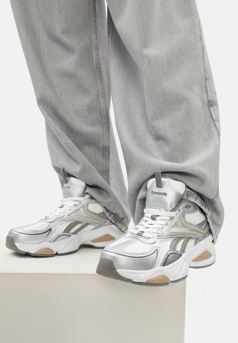 Persona ar brīviem gaiši pelēkiem džinsiem un baltiem Reebok sporta apaviem ar sudraba un bēša akcentiem, stāv uz baltas platformas.