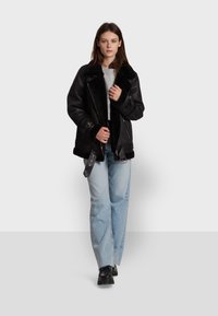 Black leather jacket with wide collar and shearling lining. Features zippers and buckle accents. Worn over a gray sweatshirt with blue jeans.