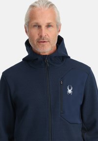Marine blauwe rits-hooded jacket van textuurstof, met een klein borstzakje en een wit spinnenlogo aan de linkerkant.