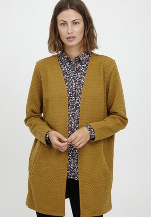Cardigan texturé jaune moutarde avec un devant ouvert, des manches longues, et une chemise à motifs floraux sombres en dessous. Un pantalon noir est porté.