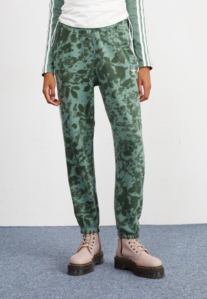 Personne portant un jogger vert à motif camouflage et des bottes beige à lacets, debout sur un sol recouvert d'un tapis bleu.