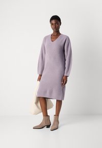 Lillāks adīts kleita ar V-veida kakla izgriezumu, garām piedurknēm un taisnu piegriezumu, savienota ar bēšiem potīšu zābakiem un krēmkrāsas plecu somu.