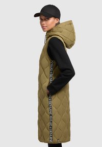 Gilet senza maniche imbottito verde oliva con cappuccio, dotato di dettagli laterali neri e strisce con logo, abbinato a una maglietta nera a maniche lunghe.