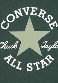 Žalias marškinėlis su baltu užrašu „CONVERSE ALL STAR“ ir „Chuck Taylor“, centre didelė blyški žalia žvaigždė. Medvilnės medžiaga, tekstūruota.