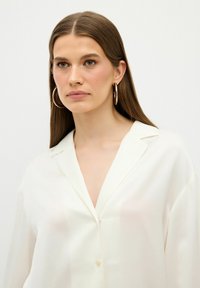 Blusa bianca leggera con colletto, caratterizzata da un scollo a V e una chiusura con bottoni. Tessuto liscio con una leggera trasparenza.