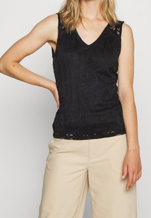 Kobieta w czarnej koronkowej bluzce bez rękawów z dekoltem w serek i beżowych szerokich spodniach, stojąca z jedną ręką na biodrze.