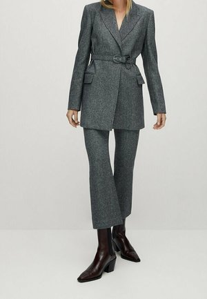 Kvinde iført en skræddersyet grå bælteblazer med matchende korte bukser og mørkebrune ankelstøvler med hæl mod en ensfarvet baggrund.
